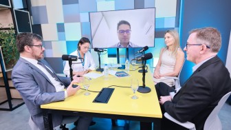 quatro pessoas sentadas &agrave; mesa, em um espa&ccedil;o retangular com decora&ccedil;&atilde;o em tons de azul e branco. No fundo uma TV exibe a imagem de uma quinta pessoa, que participa de forma virtual. 