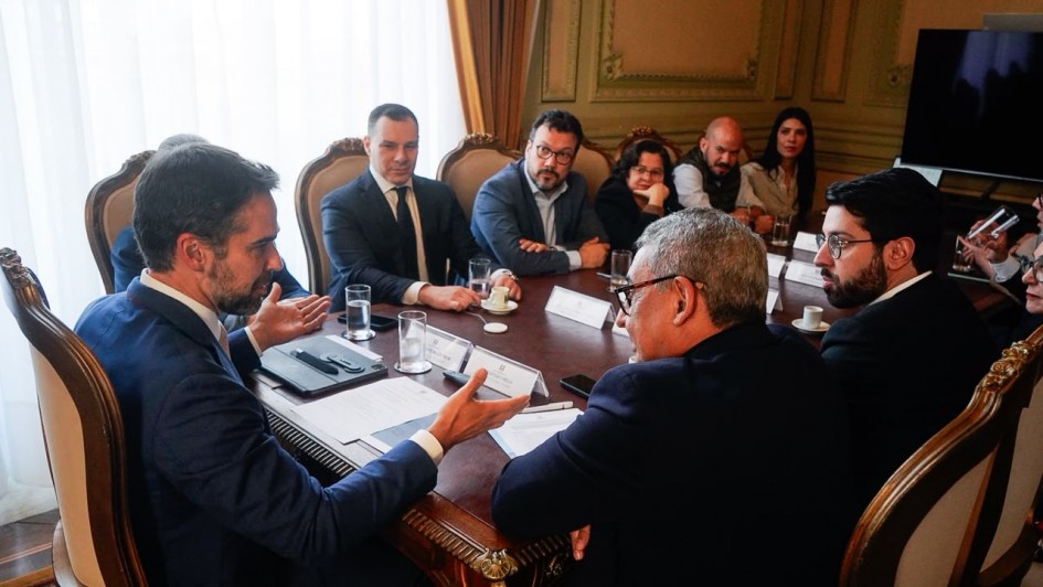 Uma fotografia colorida em plano m&eacute;dio-geral e perspectiva em &acirc;ngulo levemente elevado capta o governador Eduardo Leite reunido com gestores estaduais e autoridades ao redor de uma mesa de reuni&otilde;es no Pal&aacute;cio Piratini. **As Autoridades e a Mesa de Reuni&atilde;o (ao centro e em primeiro plano):** * &Agrave; esquerda em primeiro plano, o governador Eduardo Leite fala de perfil e gesticula com ambas as m&atilde;os abertas sobre a mesa. Ele veste um fato azul-vibrante, camisa branca e gravata clara. * &Agrave; sua frente, em primeiro plano de costas e perfil &agrave; direita, um gestor de cabelos grisalhos e &oacute;culos de arma&ccedil;&atilde;o escura escuta atentamente. * Ao seu lado, na extremidade direita, observa-se um homem jovem de barba, &oacute;culos e fato escuro. * Ao longo do lado oposto da mesa retangular de madeira nobre com cadeiras estofadas de estilo cl&aacute;ssico, alinham-se cerca de meia dezena de autoridades e secret&aacute;rios: * Destaca-se um homem de fato escuro e gravata azul-marinho, um gestor de blazer azul sobre camisa clara de bra&ccedil;os cruzados, e uma mulher de casaco escuro a apoiar o queixo na m&atilde;o. * Sobre o tampo da mesa, veem-se pl&aacute;sticas em acr&iacute;lico com identificadores nominais, pap&eacute;is de trabalho, canetas, copos de &aacute;gua e x&iacute;caras de caf&eacute;. **O Sal&atilde;o e a Ilumina&ccedil;&atilde;o (ao fundo):** * Ao fundo, as paredes em tom creme/verde-oliva suave exibem detalhes e molduras em gesso trabalhado. * &Agrave; esquerda, uma grande janela coberta por uma cortina branca transl&uacute;cida e cortinados amarelados/dourados permite a entrada de ilumina&ccedil;&atilde;o natural suave. * &Agrave; direita, ao fundo, avista-se um ecr&atilde; de televis&atilde;o desligado fixado na parede.